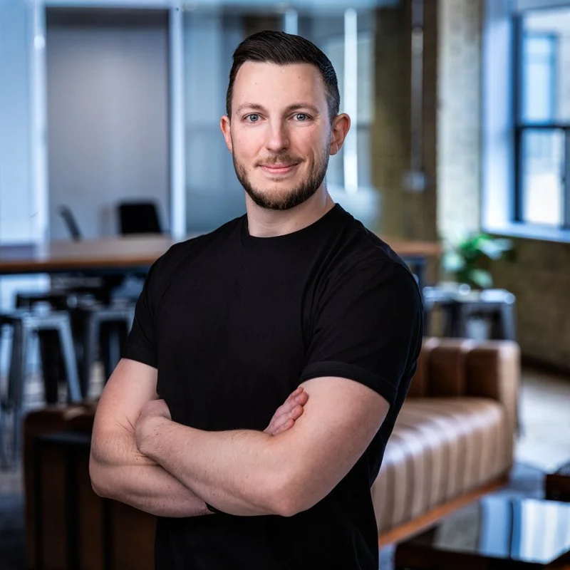 Zack O'Toole, Senior Designer and Creative Director in Toronto, short dark hair, a beard, and wearing a black t-shirt, standing in a modern, well-lit office space with arms crossed, smiling at the camera.